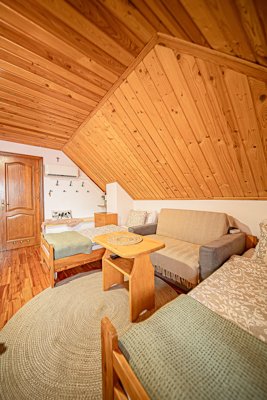 Agroturystyka Za Piecem – Przytulny pokój na poddaszu w agroturystyce ze zwierzętami, z drewnianym sufitem i ścianami, dwoma pojedynczymi łóżkami, beżową sofą, małym drewnianym stolikiem i okrągłym plecionym dywanem. Ciepłe oświetlenie i prosty, rustykalny wystrój zapewniają prawdziwy wypoczynek na wsi.