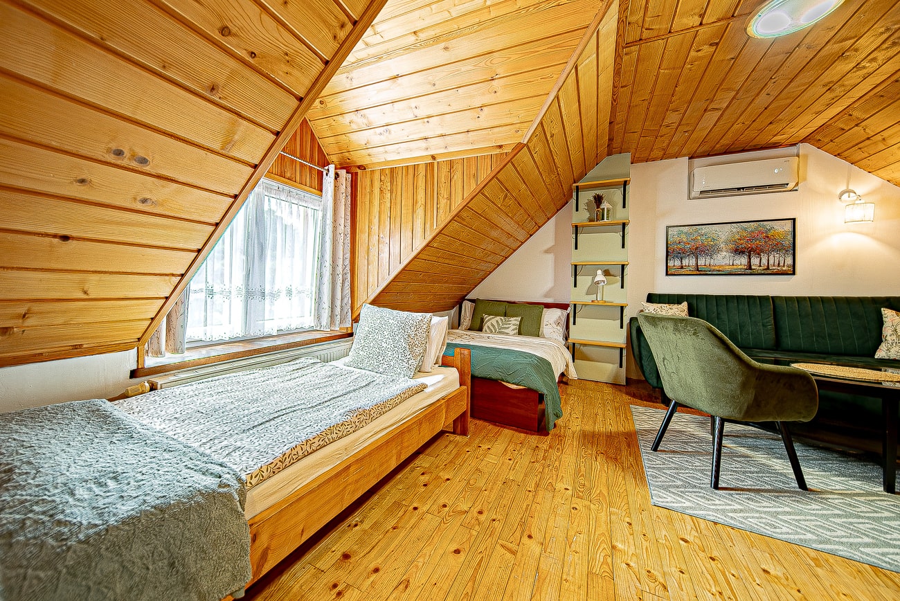 Agroturystyka Za Piecem – Przytulna sypialnia na poddaszu z drewnianymi ścianami i sufitem, dwoma pojedynczymi łóżkami, zieloną sofą, małym stolikiem z krzesłami, oknem z zasłonami, półką na książki, klimatyzatorem i grafiką na ścianie. Ciepłe oświetlenie tworzy przyjazną atmosferę dla wypoczynku na wsi.