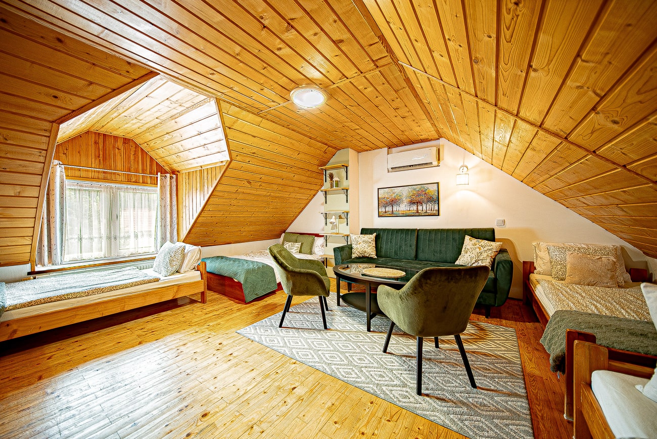 Agroturystyka Za Piecem – Przytulny pokój na poddaszu z drewnianym sufitem i podłogą, wyposażony w dwa pojedyncze łóżka, zieloną sofę, dwa krzesła, mały okrągły stolik, półki na książki, dywan i duże okna - idealny na agroturystykę z dziećmi lub wypoczynek na wsi.