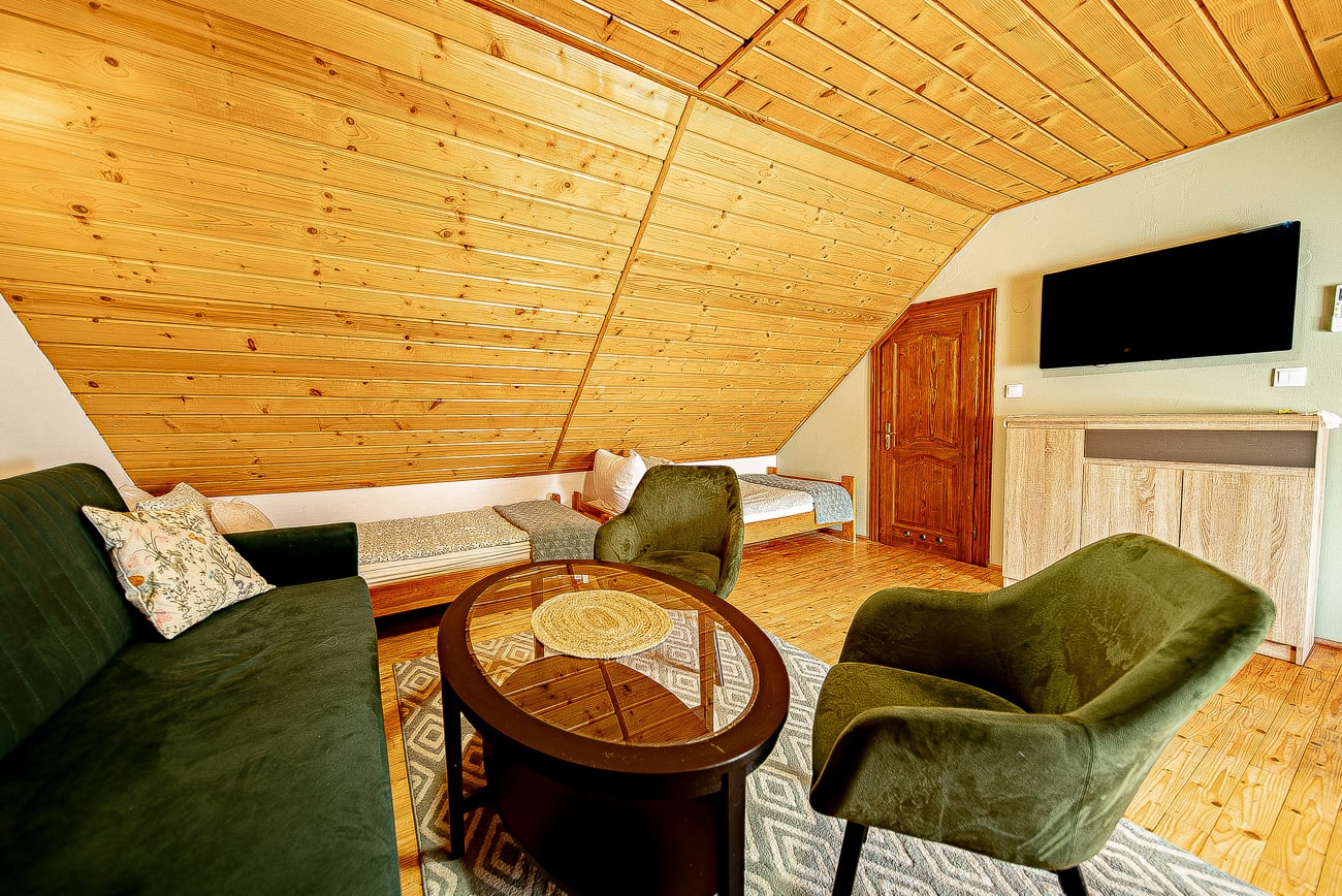 Agroturystyka Za Piecem – Przytulny pokój na poddaszu ze skośnym drewnianym sufitem, zielonymi fotelami, ciemną sofą, okrągłym stolikiem kawowym, dwoma pojedynczymi łóżkami - idealny na wypoczynek na wsi z rodziną - wzorzystym dywanem, drewnianymi drzwiami i zamontowanym na ścianie telewizorem nad szafką z jasnego drewna.