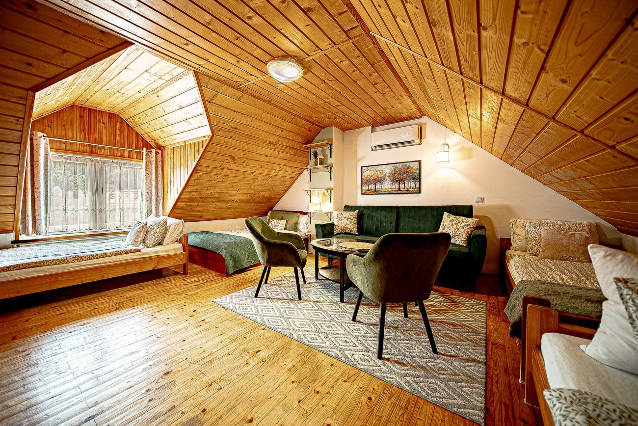 Agroturystyka Za Piecem – Przytulny pokój na poddaszu ze skośnym drewnianym sufitem, łóżkiem przy dużym oknie, zieloną sofą i fotelami wokół małego stolika. Idealny na wypoczynek na wsi, z ciepłym oświetleniem i wzorzystym dywanem na wypolerowanej drewnianej podłodze.