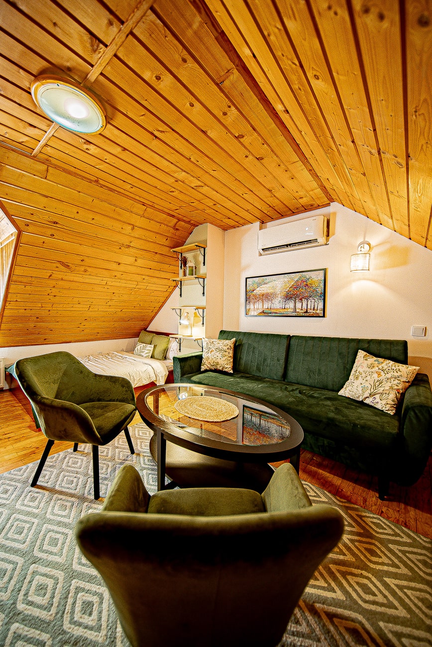 Agroturystyka Za Piecem – Przytulny salon na poddaszu ze skośnym drewnianym sufitem, zielonymi sofami i fotelami, okrągłym stolikiem kawowym, dekoracyjnymi poduszkami, grafiką na ścianie, klimatyzacją i wzorzystym dywanem. Idealny na noclegi dla rodzin z dziećmi szukających wypoczynku na wsi. Łóżko w narożniku.