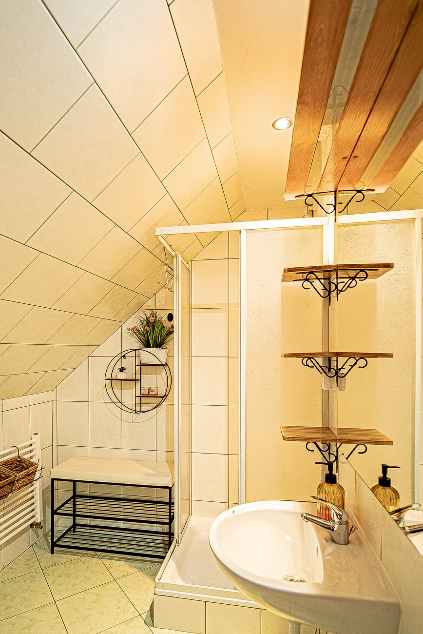 Agroturystyka Za Piecem – Mała, nowoczesna łazienka z białymi kafelkami na ścianach i podłodze, szklaną kabiną prysznicową, umywalką z lustrem, drewnianymi półkami, ławką i ozdobnymi roślinami - idealna na agroturystykę z dziećmi lub noclegi dla rodzin z dziećmi.