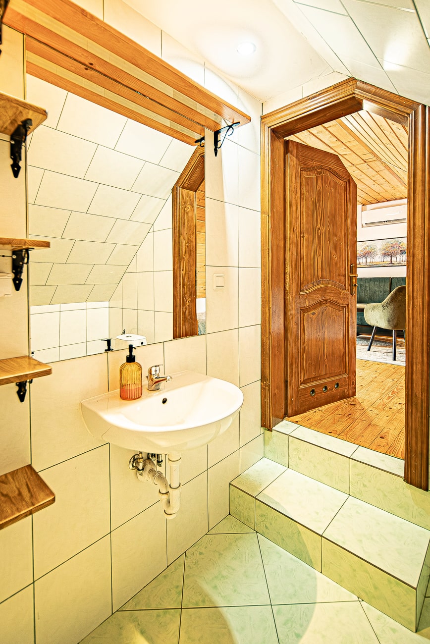 Agroturystyka Za Piecem – Mała łazienka wyłożona kafelkami z białą umywalką, lustrem i drewnianymi półkami. Schody prowadzą do otwartych drewnianych drzwi odsłaniających wyłożony drewnem pokój z krzesłem - idealny na wypoczynek na wsi lub agroturystykę z dziećmi.