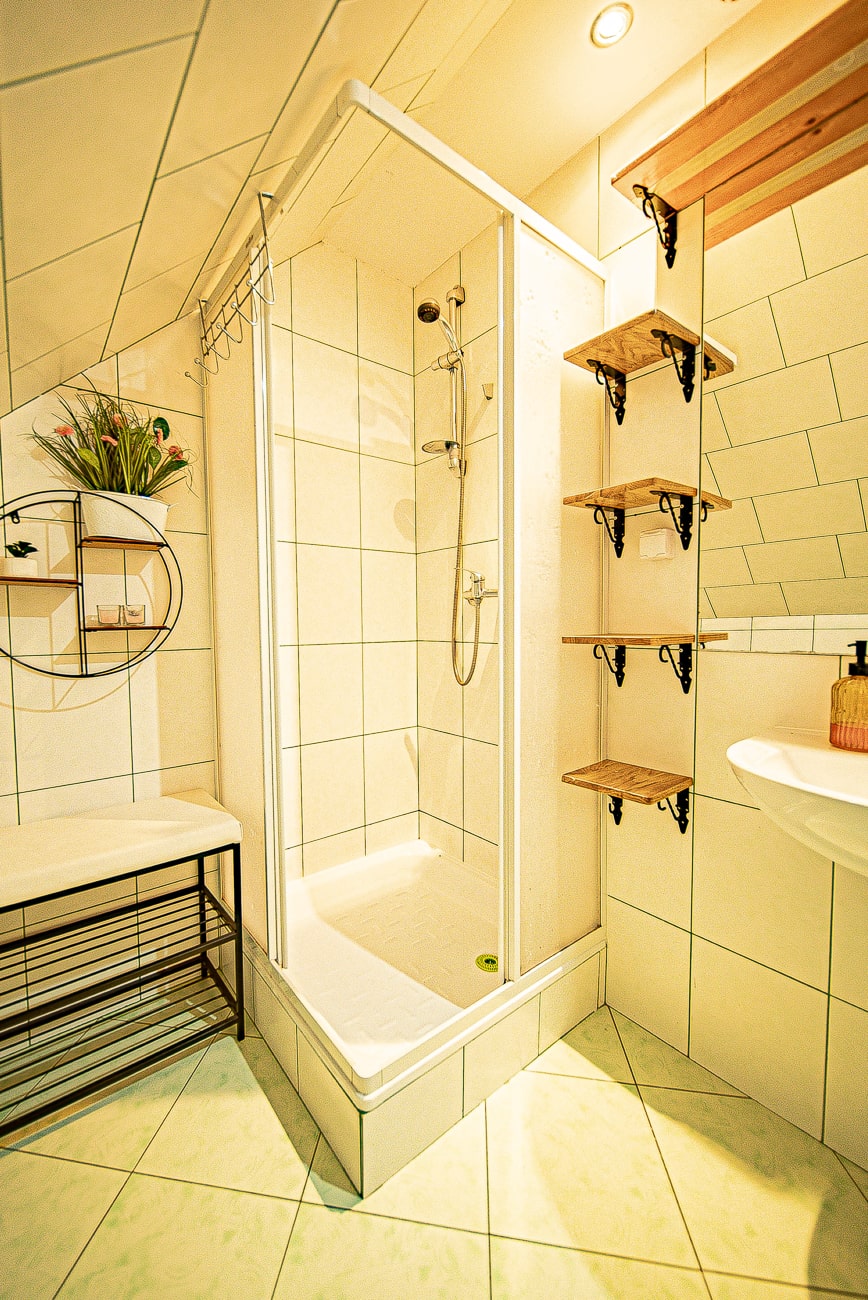 Agroturystyka Za Piecem – Mała, nowoczesna łazienka w spa na wsi ma szklany prysznic, białe kafelki na ścianach i podłodze, drewniane półki, ławkę i rośliny doniczkowe dodające dekoracyjnych akcentów.