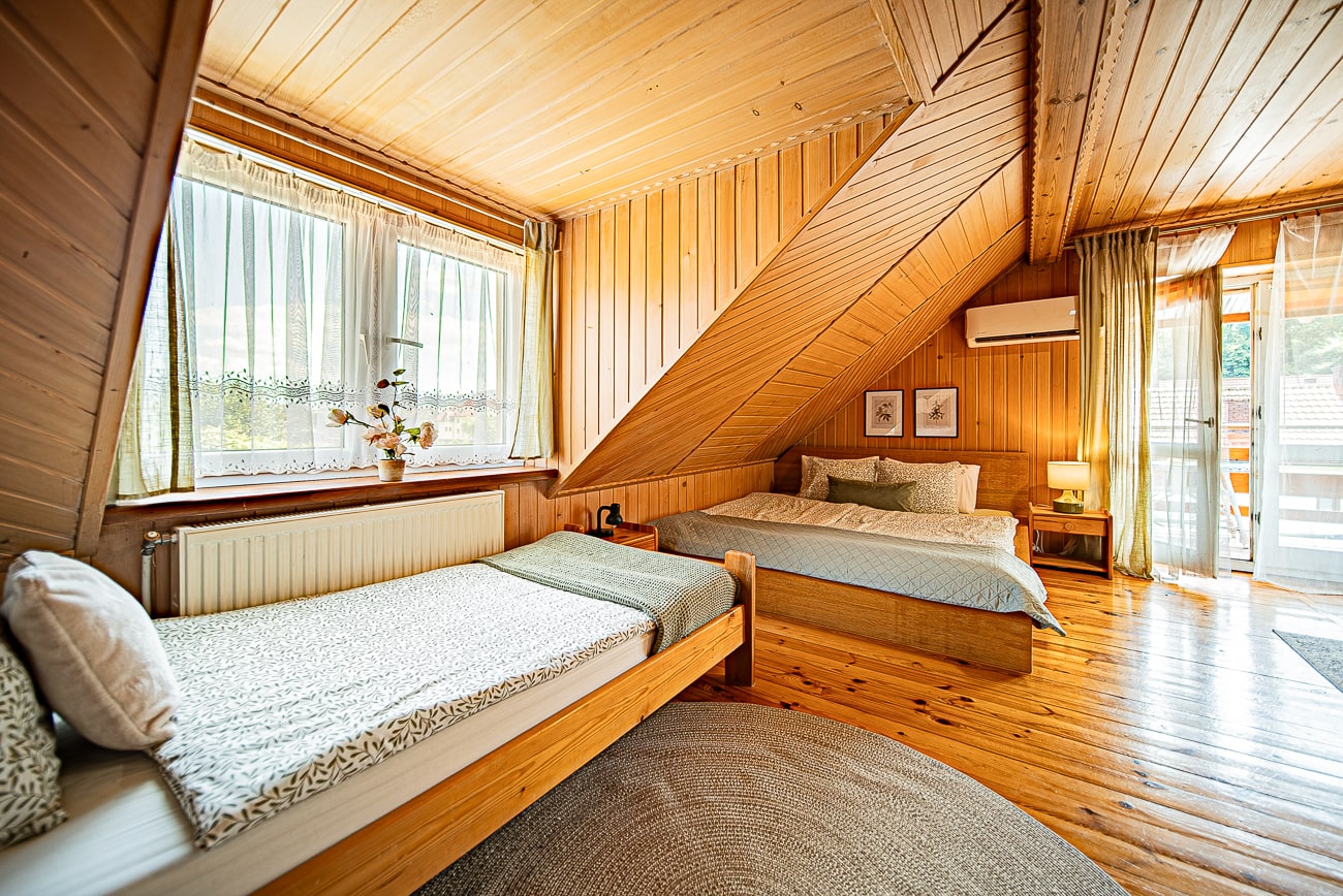 Agroturystyka Za Piecem – Przytulna sypialnia na poddaszu z drewnianymi panelami na ścianach i suficie, pojedynczymi i podwójnymi łóżkami, zielonymi akcentami, oknem z zasłonami, oprawionymi dziełami sztuki, szafką nocną i drzwiami na balkon - idealna na agroturystykę dla rodziny lub spa na wsi.