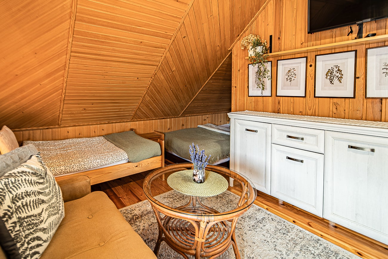 Agroturystyka Za Piecem – Przytulny pokój na poddaszu z drewnianymi panelami na ścianach i suficie, dwoma pojedynczymi łóżkami, białą komodą z oprawionymi botanicznymi nadrukami, rattanowym stolikiem kawowym z lawendowym wazonem i beżową sofą - idealny na agroturystykę dla rodziny lub wypoczynek z dziećmi.