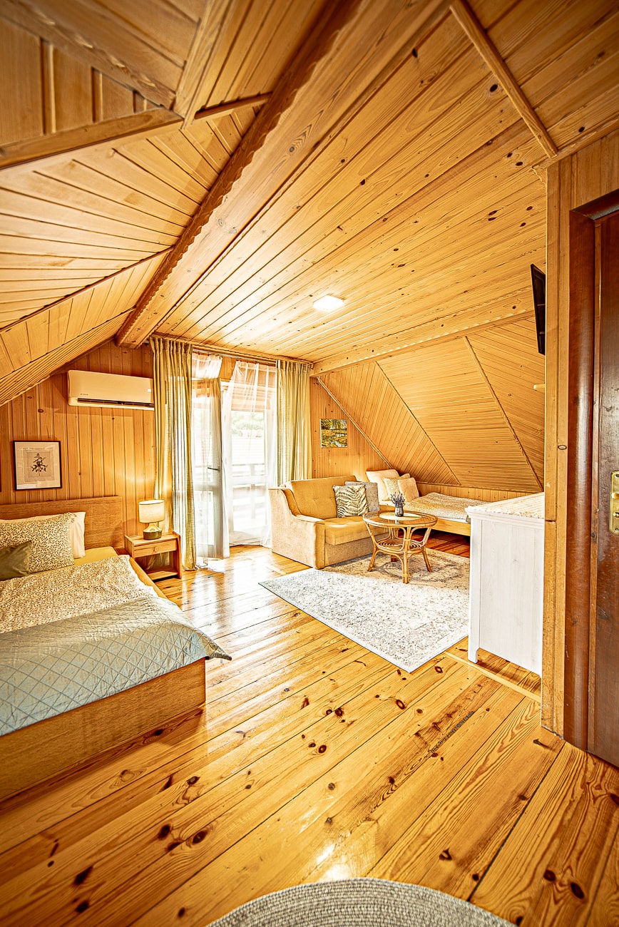 Agroturystyka Za Piecem – Przytulny pokój na poddaszu z drewnianymi ścianami i sufitem, łóżkiem, sofą, małym stolikiem i dywanem. Naturalne światło wpada przez szklane drzwi z zasłonami, tworząc ciepłą atmosferę - idealną na agroturystykę dla rodziny lub wypoczynek na wsi.