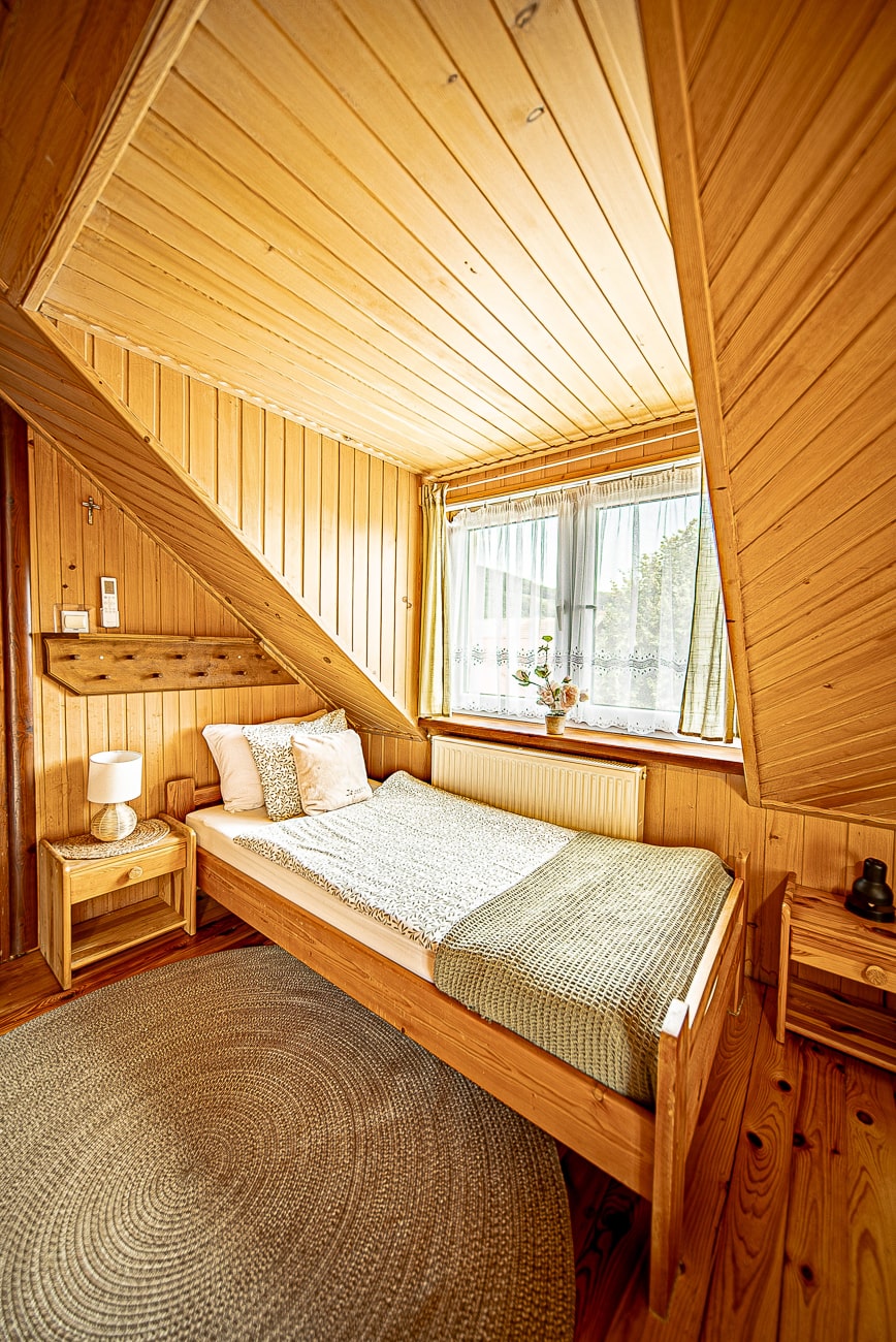 Agroturystyka Za Piecem – Przytulna sypialnia na wypoczynek na wsi, z drewnianymi ścianami i sufitem, starannie wykonanym łóżkiem w kolorze białym i zielonym, okrągłym dywanikiem, szafkami nocnymi z lampkami i oknem wypełniającym przestrzeń naturalnym światłem.