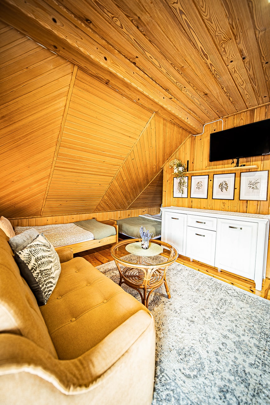 Agroturystyka Za Piecem – Przytulny pokój na poddaszu ze skośnym drewnianym sufitem, beżową sofą, małym łóżkiem, okrągłym szklanym stolikiem kawowym, białą szafką, telewizorem na ścianie, dziełami sztuki oprawionymi w ramki i szarym wzorzystym dywanem. Idealny do agroturystyki z dziećmi w jasnej i zachęcającej atmosferze.