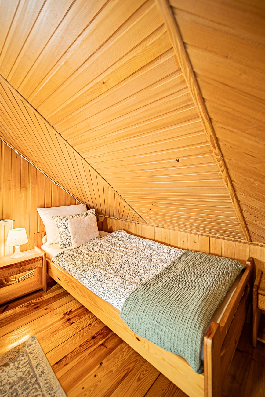 Agroturystyka Za Piecem – Przytulna drewniana sypialnia na poddaszu w agroturystyce z dziećmi wyposażona jest w pojedyncze łóżko z wzorzystą pościelą, zielony koc, puszyste poduszki, szafkę nocną z lampką oraz ciepłe drewniane panele na ścianach i suficie.