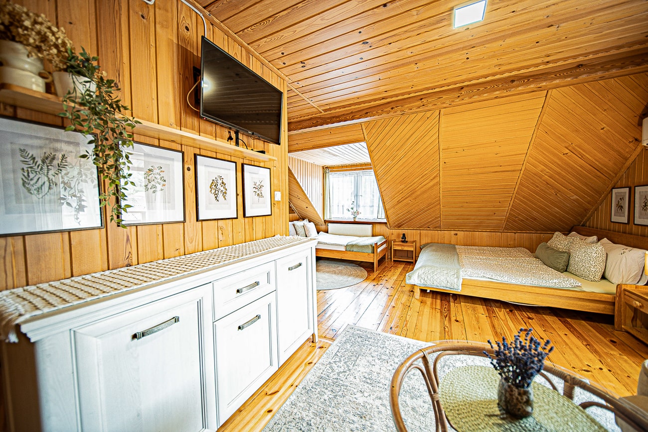 Agroturystyka Za Piecem – Przytulna sypialnia na poddaszu z drewnianymi ścianami i sufitem, podwójnym łóżkiem, pojedynczym łóżkiem przy oknie, białymi meblami, oprawionymi botanicznymi nadrukami, telewizorem z płaskim ekranem i małym okrągłym stolikiem z wazonem lawendy - idealna na noclegi dla rodzin z dziećmi szukającymi wypoczynku na wsi.