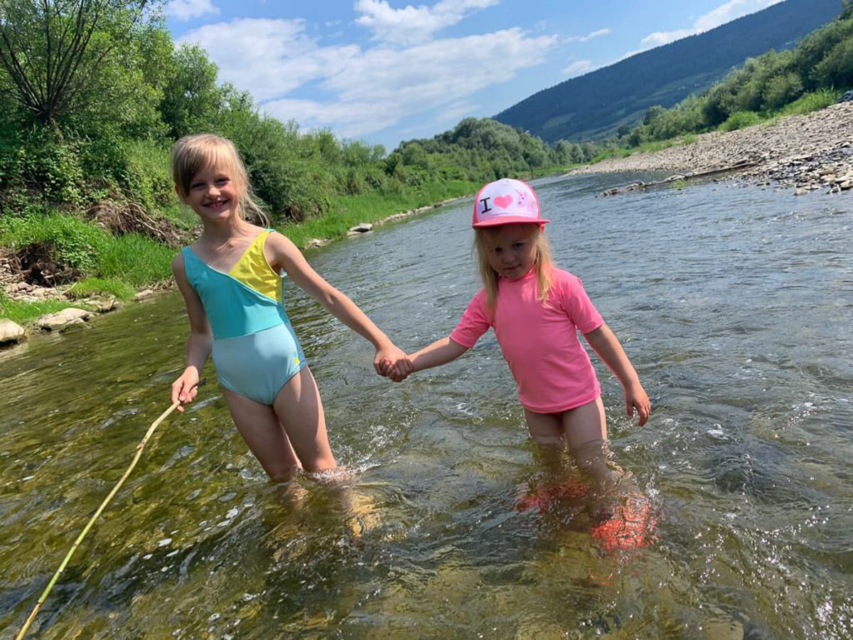 Agroturystyka Za Piecem – Dwie młode dziewczyny stoją trzymając się za ręce w płytkim, skalistym strumieniu otoczonym zielenią i wzgórzami, ciesząc się wypoczynkiem na wsi. Jedna ma na sobie strój kąpielowy i trzyma kij, podczas gdy druga jest w różowym stroju z czapką pod częściowo zachmurzonym niebem.