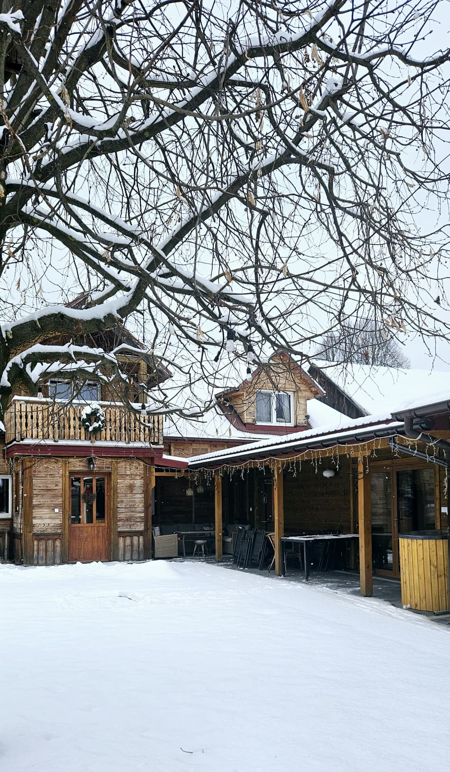 Agroturystyka Za Piecem – Drewniany dom z balkonem i zadaszonym patio stoi pod dużym, bezlistnym drzewem. Śnieg pokrywa ziemię i dachy, tworząc przytulną agroturystykę dla rodzin, idealną na zimowe pobyty.