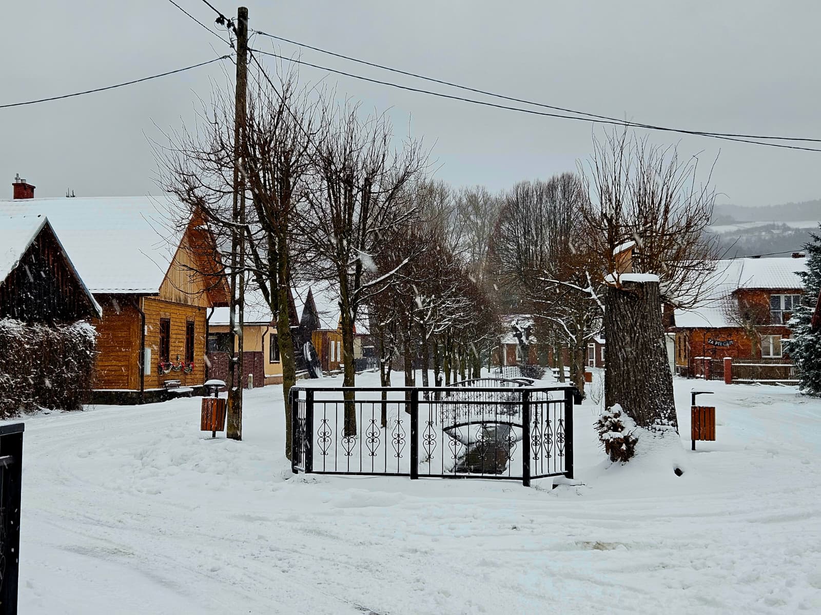 Agroturystyka Za Piecem – Ośnieżona wiejska ulica z gołymi drzewami, drewnianymi domami i czarną metalową bramą to sceneria dla agroturystyki z dziećmi. Śnieg pokrywa ziemię, dachy i gałęzie w pochmurny zimowy dzień - idealny na spokojny wypoczynek na wsi.