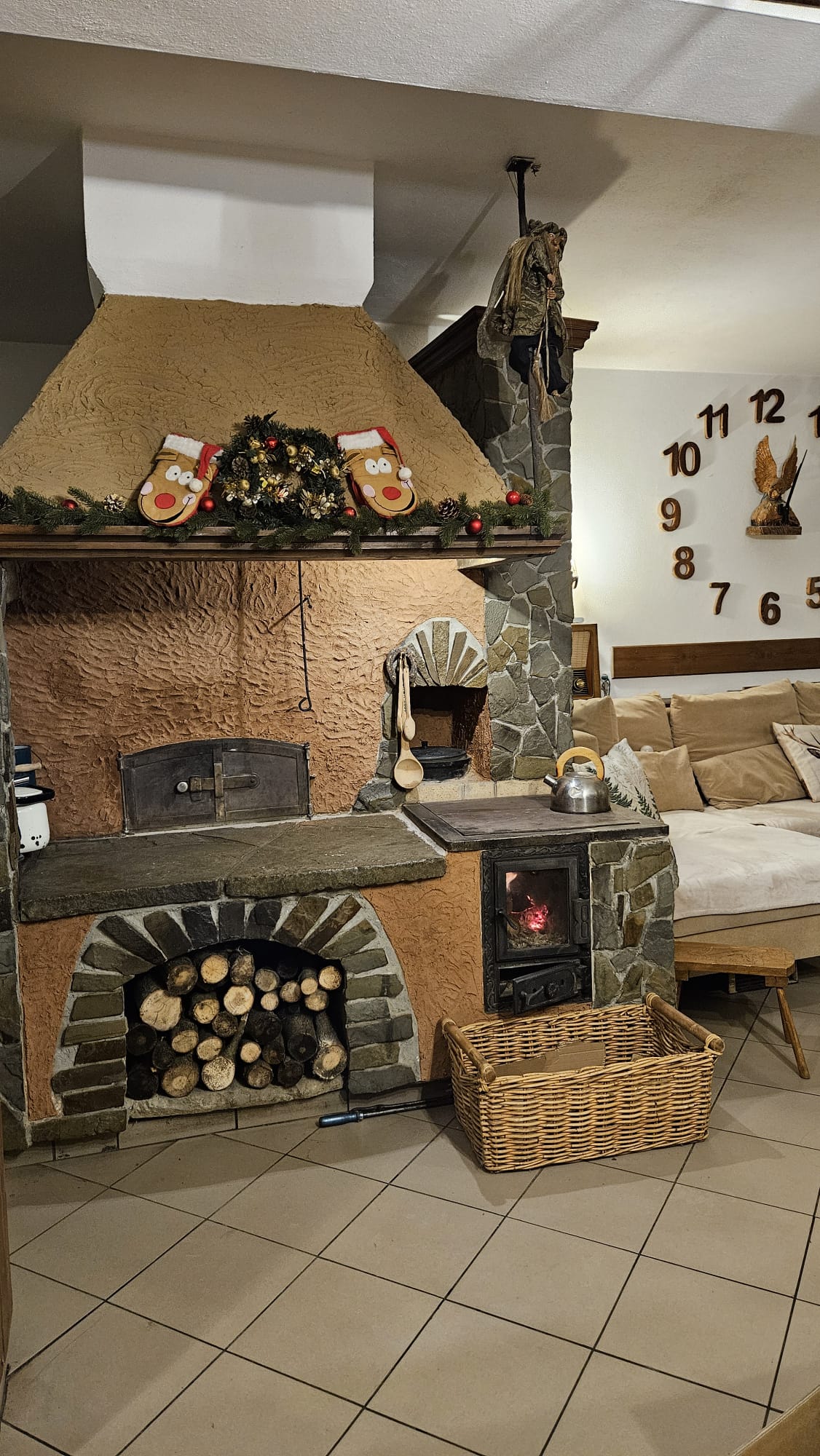 Agroturystyka Za Piecem – Rustykalny kamienny kominek z piecem opalanym drewnem, drewno opałowe ułożone poniżej, mała kuchenka z czajnikiem oraz świąteczne pończochy i wieniec powyżej. Idealny do agroturystyki dla rodziny - ciesz się przytulnymi chwilami na kanapie pod dużym zegarem ściennym.