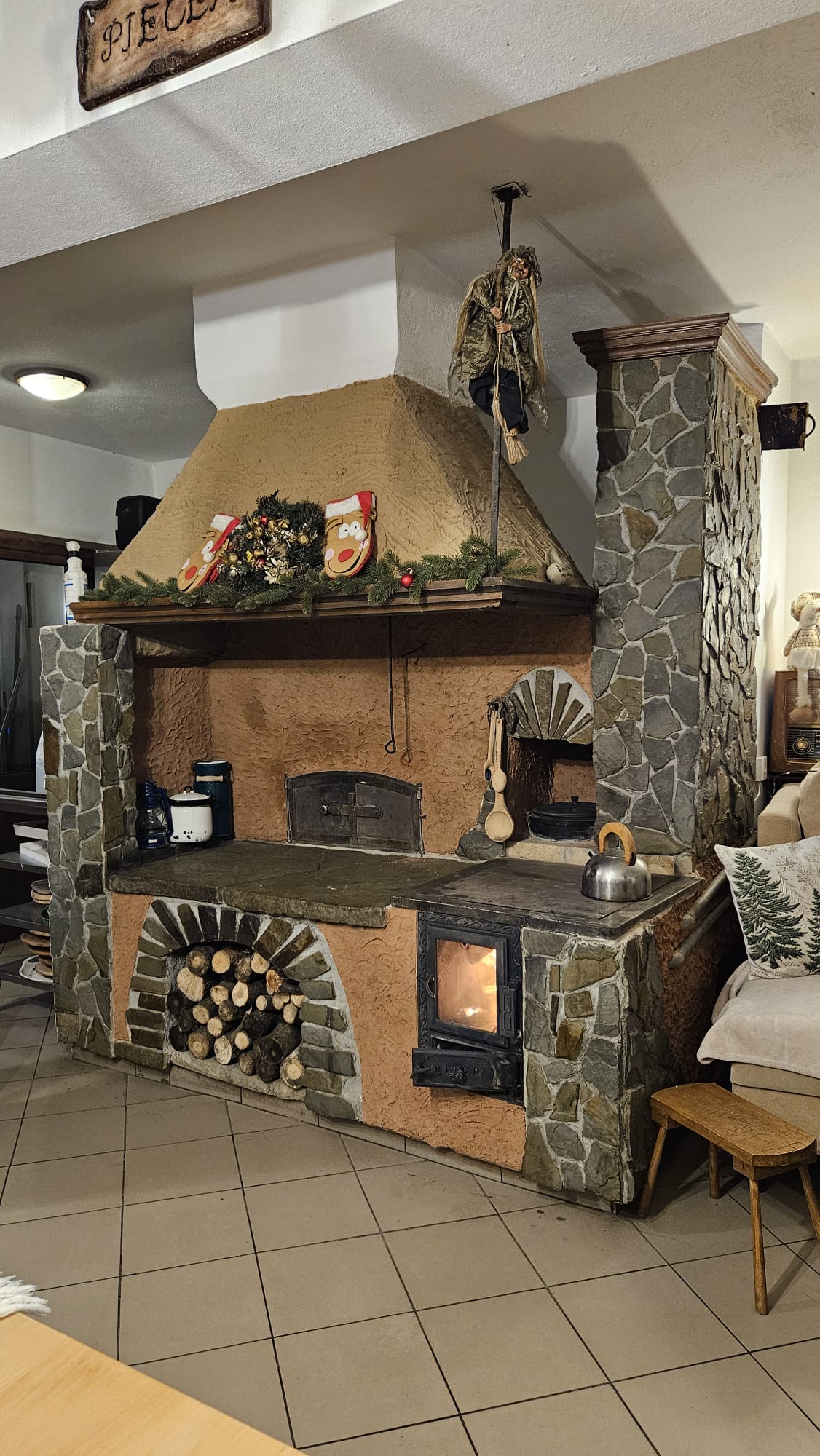 Agroturystyka Za Piecem – Rustykalny kominek i piec opalany drewnem z kamiennym wykończeniem i sztukaterią, udekorowany na Boże Narodzenie, otoczony przytulnymi meblami - idealny dla agroturystyki dla rodzin poszukujących ciepła i wiejskiego uroku.