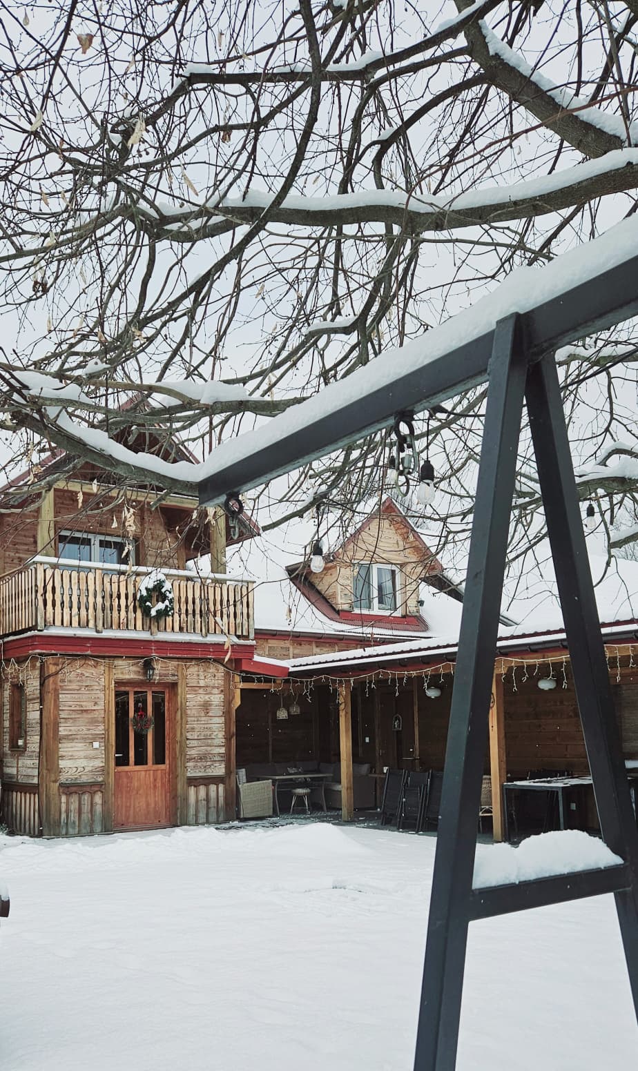 Agroturystyka Za Piecem – Pokryta śniegiem huśtawka czeka przed rustykalnym drewnianym domem z balkonem, otoczonym zimowym uściskiem - idealnym na wypoczynek na wsi lub noclegi dla rodzin z dziećmi szukających spokojnego wiejskiego uroku.