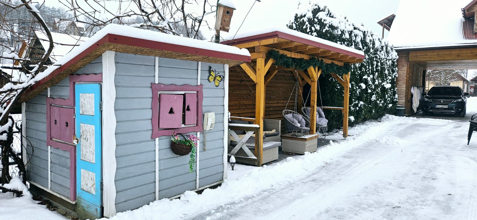 Agroturystyka Za Piecem – Mały, kolorowy drewniany domek z różowymi oknami i niebieskimi drzwiami stoi na zaśnieżonym podwórku, idealny do agroturystyki z dziećmi. Nieopodal znajduje się drewniana weranda z krzesłami, a dalej na zaśnieżonym podjeździe zaparkowany jest czarny SUV.