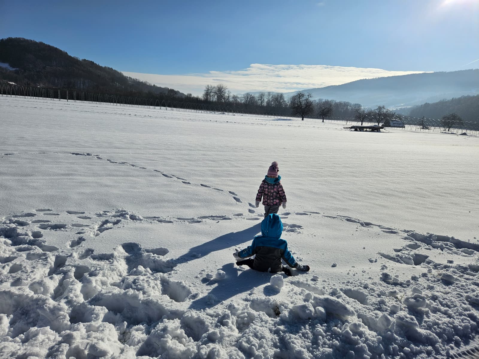 Agroturystyka Za Piecem – Dwoje dzieci bawi się na zaśnieżonym polu pod czystym, błękitnym niebem, jedno stoi, a drugie siedzi na śniegu. Ślady stóp prowadzą w kierunku odległych drzew i wzgórz, oddając radość rodzinnego wypoczynku na wsi.