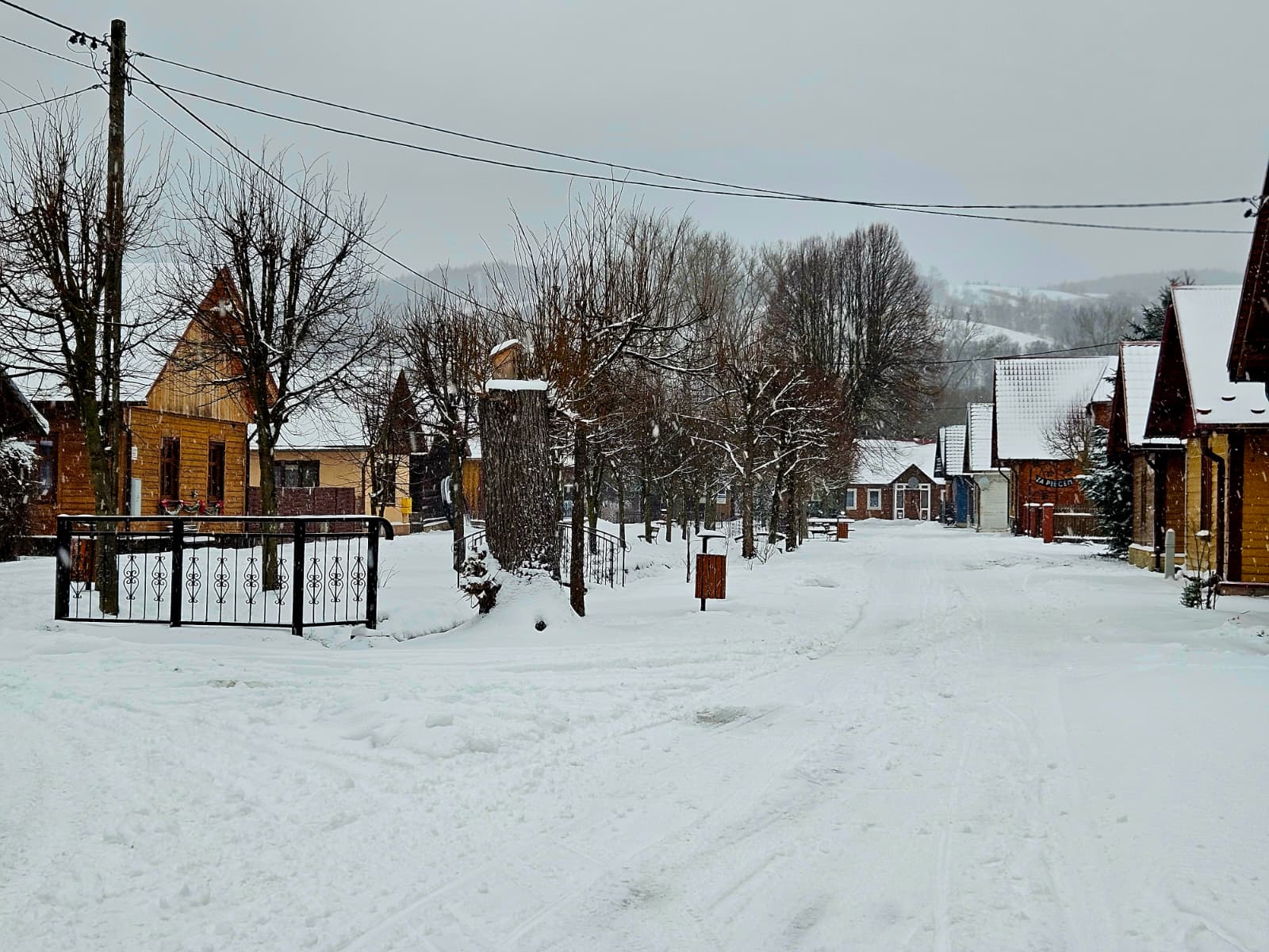 Agroturystyka Za Piecem – Ośnieżona wiejska ulica wyłożona drewnianymi domami i nagimi drzewami, idealna na agroturystykę z dziećmi. Śnieg pokrywa ziemię i dachy, a wzgórza i bezlistne drzewa rozciągają się na zachmurzonym niebie.