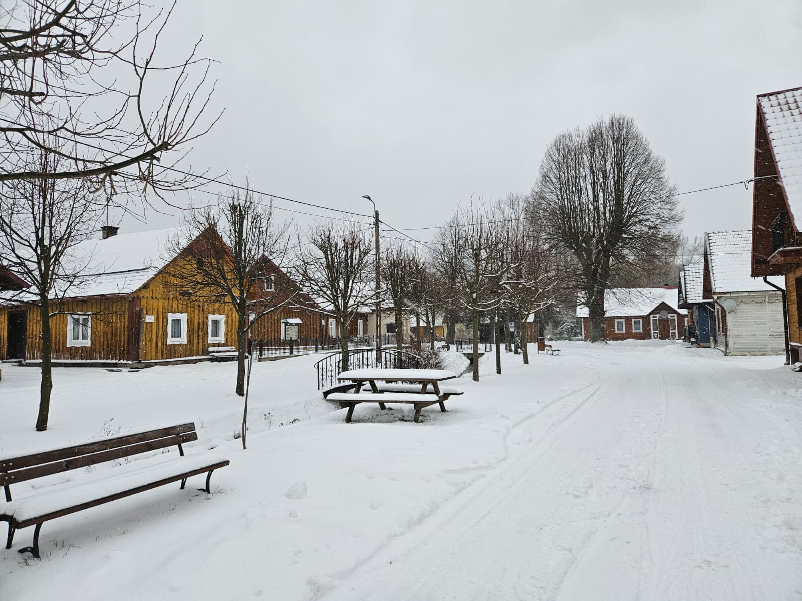 Agroturystyka Za Piecem – Ośnieżona wiejska sceneria z drewnianymi domami, nagimi drzewami, ławkami, stołem piknikowym i śniegiem pokrywającym ziemię pod zachmurzonym niebem - idealne miejsce na agroturystykę z dziećmi lub relaksujące spa na wsi.