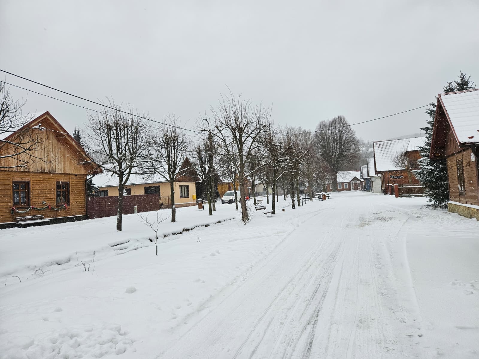 Agroturystyka Za Piecem – Cicha wiejska ulica pokryta śniegiem, wyłożona drewnianymi domami i bezlistnymi drzewami. Niebo jest zachmurzone, a sceneria wygląda na spokojną i zimową - idealną dla tych, którzy szukają wypoczynku na wsi lub agroturystyki dla rodziny.