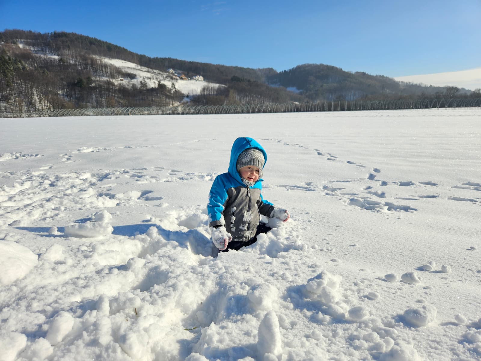Agroturystyka Za Piecem – Małe dziecko w niebieskiej zimowej kurtce i szarej czapce siedzi w głębokim śniegu, trzymając śnieg w obu rękach, otoczone zaśnieżonymi wzgórzami i drzewami - idealna sceneria dla agroturystyki dla rodzin, które chcą wspólnie cieszyć się przyrodą.