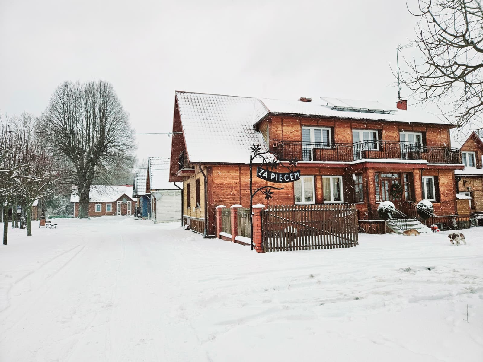 Agroturystyka Za Piecem – Drewniany dom z napisem ZA PIECEM stoi na zaśnieżonej ulicy, idealnej na agroturystykę dla rodziny. Drzewa i płoty pokryte są śniegiem, a w tle pod zachmurzonym zimowym niebem widać kolejny budynek.