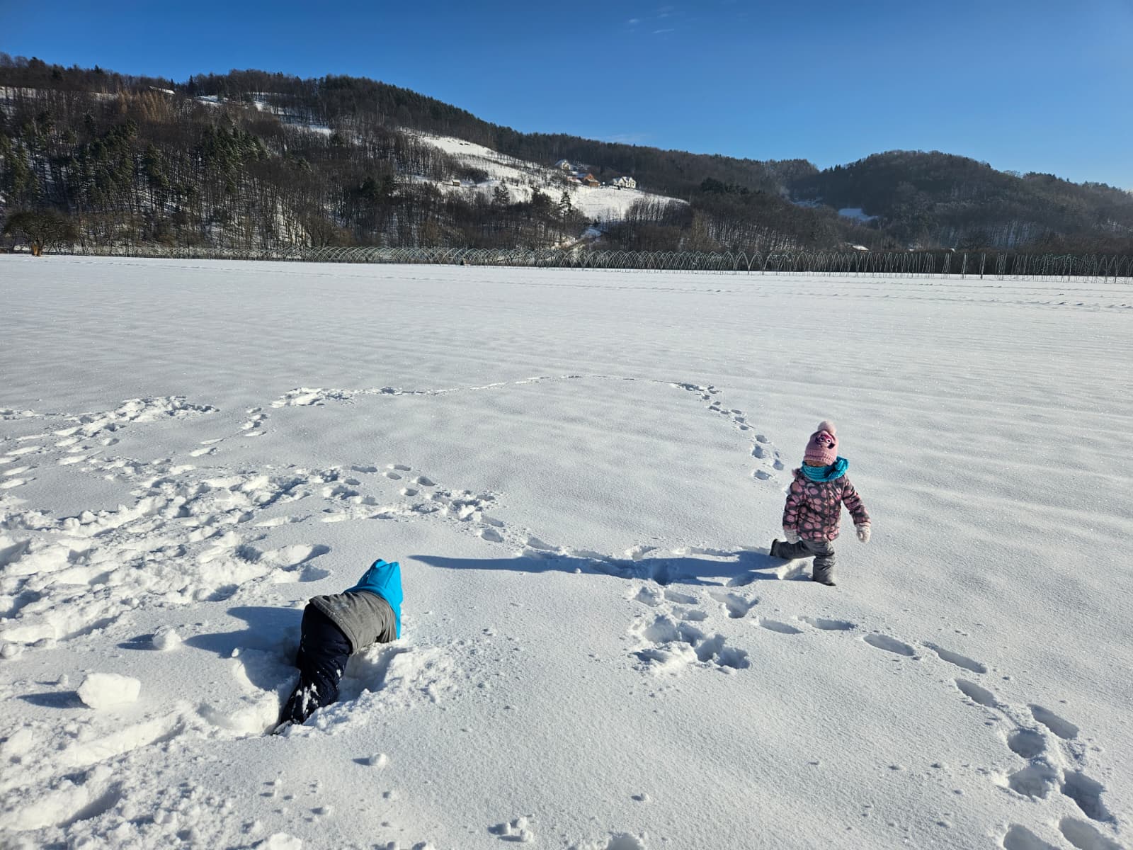 Agroturystyka Za Piecem – Dwoje małych dzieci cieszy się agroturystyką z dziećmi, bawiąc się na zaśnieżonym polu pod czystym, błękitnym niebem. Jedno z nich klęczy na śniegu, podczas gdy drugie stoi obok, a w tle widać ośnieżone wzgórza i drzewa.