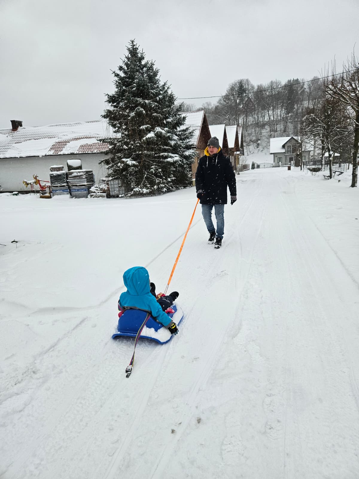 Agroturystyka Za Piecem – Dorosły ciągnie dziecko na niebieskich sankach po zaśnieżonej wiejskiej drodze, otoczonej ośnieżonymi domami, drzewami i wzgórzami - idealne miejsce na agroturystykę z dziećmi i wspólną zimową zabawę na wsi.